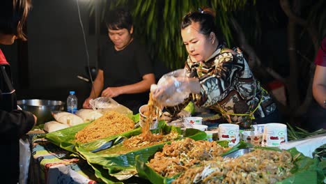 Pad-Thai,-Berühmtes-Thailändisches-Essen,-Verkäufer-Auf-Einem-Nachtmarkt,-Die-Damit-Beschäftigt-Sind,-Einen-Müden-Kunden-Nach-Der-Arbeit-Zu-Bedienen