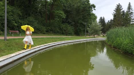 Woman-in-white-dress,-yellow-umbrella-walks-near-lake-with-reflection