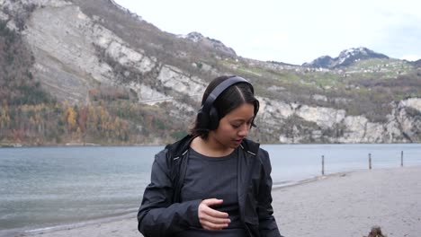 Young-woman-sits-on-the-shore-of-a-mountain-lake-and-puts-on-her-headphones--Tracking-shot