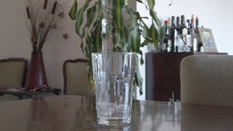 pouring-water-vertically-into-a-glass-with-a-background-of-a-livingroom,-in-slowmotion
