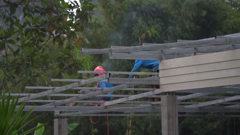 Thai-Welders-on-steel-roof-beams-doing-their-job-without-safety-equipment,-wide-shot-zoom-to-close-up-shot-in-slow-motion