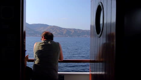 Vista-Desde-Detrás-De-Un-Macho-Adulto-Desde-El-Interior-De-La-Cabina-Apoyándose-En-La-Barandilla-Del-Barco-Mientras-Cruza-El-Mar-Jónico