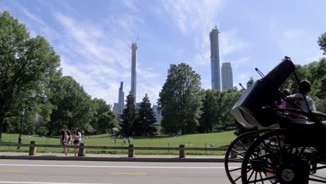 View-of-skyscrapers-under-construction-in-Manhattan-from-Central-Park,-with-a-Horse-Carriage,-Rikshas,-runners-and-tourists-passing-by-in-the-foreground-at-Terrace-Drive,-New-York-City