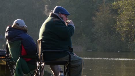 Retired-pensioner-gentleman-sitting-in-autumn-park-drinking-warm-steaming-beverage-from-flask-looking-out-across-park-lake