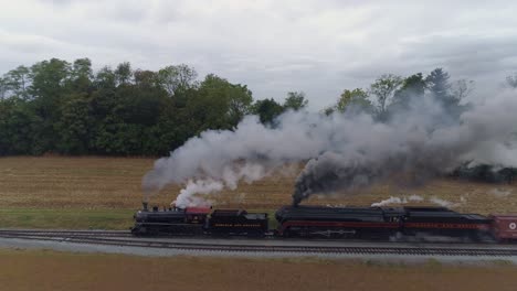 Vista-Lateral-Aérea-De-Dos-Locomotoras-De-Vapor-Que-Se-Dirigen-Dos-Veces-A-Un-Tren-De-Carga-Que-Sopla-Junto-Con-Humo-Negro-Y-Vapor