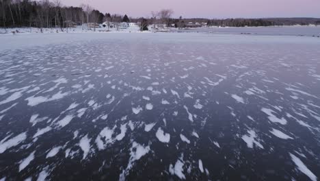 Flying-low-towards-shore-above-snowy-patterns-in-dark-ice-at-dawn-AERIAL
