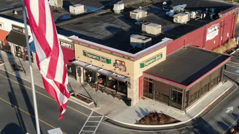 Aerial-turn-reveals-Primanti-Bros,-Hair-Cuttery,-Inside-Track-stores-under-American-flag