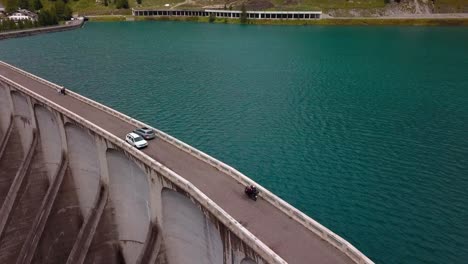 Presa-Del-Lago-Fedaia-En-Las-Montañas-Dolomitas-Con-Un-Coche-Gris-Y-Una-Motocicleta-Pasando-La-Carretera-Por-Encima-De-La-Estructura-De-La-Pared,-Tiro-De-Seguimiento-De-La-órbita-Aérea-De-Drones