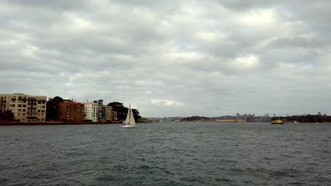 Sailboat-on-the-harbor-with-apartment-buildings-and-seaplane-far-in-the-sky-on-an-overcast-day,-Stable-handheld-shot