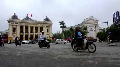 Hanoi-Opera-House-Und-Hanoi-Hilton-Mit-Starkem-Verkehr-Von-Motorrädern-Und-Autos,-Die-Am-Platz-Der-Augustrevolution-Vorbeifahren,-Niedriger-Winkel,-Gesperrter-Schuss