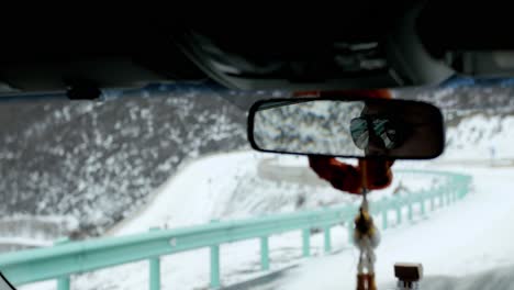 Rear-view-mirror-driving-snowy-Chinese-mountain-roads-alongside-barrier,-reflecting-asphalt-pathway-behind