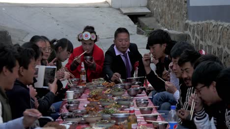 A-Chinese-newly-wed-couple-sitting-at-the-end-of-a-long-table-in-Weixi,-Yunnan,-eating-with-friends-and-family---Zoom-in