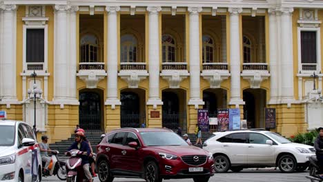 Entrada-De-La-ópera-De-Hanoi-Con-Tráfico-Denso-De-Motos-Y-Automóviles-Que-Pasan-Por-La-Plaza-De-La-Revolución-De-Agosto,-Toma-De-Mano