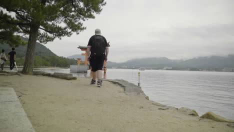 Kaukasischer-Rucksacktourist-Geht-In-Richtung-Heiliges-Japanisches-Tori-Tor-Am-Meer-Auf-Der-Insel-Itsukushima,-Miyajima,-Japan