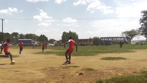 Una-Toma-En-Cámara-Lenta-De-Jóvenes-Africanos-Jugando-Al-Fútbol-En-Un-Campo-De-Tierra-Al-Lado-De-Una-Carretera