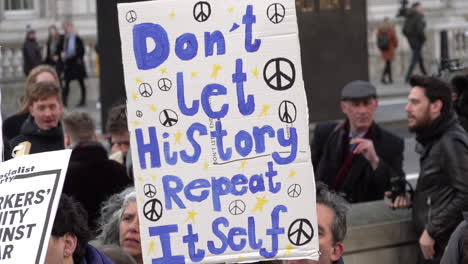 Una-Persona-En-Una-Protesta-Que-Se-Opone-A-La-Guerra-Con-Irán-Sostiene-Una-Pancarta-Cubierta-Con-Símbolos-De-Paz-Y-Una-Escritura-Azul-Que-Dice-&quot;no-Dejes-Que-La-Historia-Se-Repita&quot;