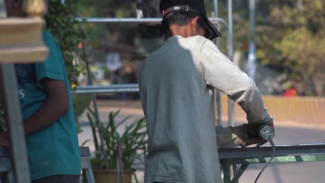 Close-Slow-Motion-Shot-of-an-Asian-Man-at-the-Side-of-the-Road-Using-an-Angle-Grinder-on-a-Piece-of-Metal