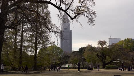 Leute,-Die-Im-Japanischen-Garten-Shinjuku-Gyoen-In-Tokio,-Japan,-Mit-Grünen-Bäumen-In-Der-Nähe-Von-Hochhausturm-Und-Gebäude-Spazieren-Gehen---Breite-Aufnahme