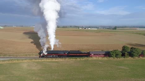 Vista-Aérea-De-Lado-A-Lado-De-Un-Motor-De-Vapor-Nw-Soplando-Vapor-En-El-Campo-Preparándose-Para-Tirar-De-Automóviles-De-Pasajeros-Con-Vista-De-Las-Tierras-De-Cultivo