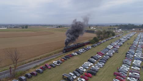 Luftwinkelansicht-Einer-Neuen-Dampfmaschine,-Die-Dampf-Und-Rauch-Bläst,-Während-Sie-An-Einem-Sonnigen-Tag-Personenkraftwagen-Mit-Blick-Auf-Geparkte-Autos-Zieht