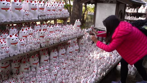 Chica-Con-Chaqueta-Rosa-Tomando-Fotos-De-Maneki-neko,-También-Conocido-Como-Gatos-De-La-Suerte-O-Estatua-De-Gatos-Que-Llaman-Dentro-Del-Templo-Gotokuji-En-Tokio,-Japón