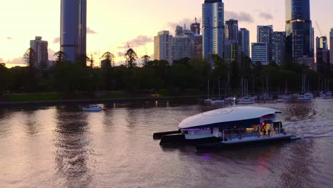 Luxurious-Yacht-Entertainment-Venue-Passing-Across-The-Serene-Water-With-Modern-Towers-And-Buildings-Cityscape-During-Sundown-In-Brisbane,-Australia