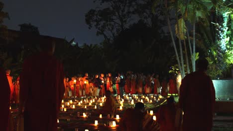 Wide-Exterior-Shot-of-Monks-Walking-in-a-Line-With-Candles-Lit-in-the-Mid-Ground-and-a-Few-Monks-Standing-and-Sitting-in-the-Fore-Ground-in-a-Ceremony-With-a-Camera-Man-Filming-in-the-Night-Time