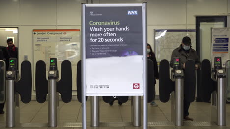 People-in-protective-surgical-face-masks-enter-and-exit-an-underground-station-and-pass-an-NHS-health-sign-waring-the-to-public-to-wash-hands-for-twenty-seconds-during-the-Coronavirus-outbreak