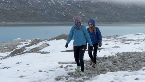 Two-Women-Hiking-on-a-Snow-Rocky-Landscape-in-Front-of-a-Colorful-Glacial-Lake-in-Norway,-Slow-Motion