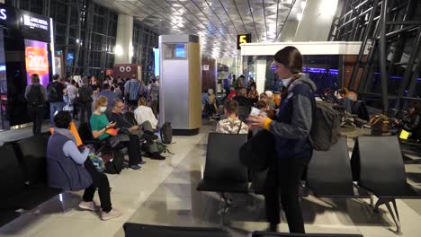 Cautious-travelers-with-face-masks-waiting-on-the-airport,-man-and-woman-on-the-seats-in-front-of-a-terminal-waiting-for-their-flight-to-get-home