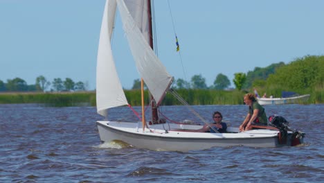 Two-young-guys-in-sailboat-waving-and-pointing-at-camera-on-blue-lake-on-sunny-day