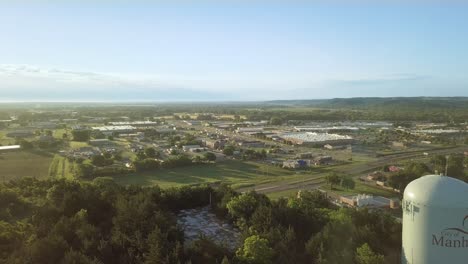 Tall-steel-water-tower-at-plateau-of-green-rural-hill-in-countryside-with-view-of-city-and-sun-flare,-backward-aerial