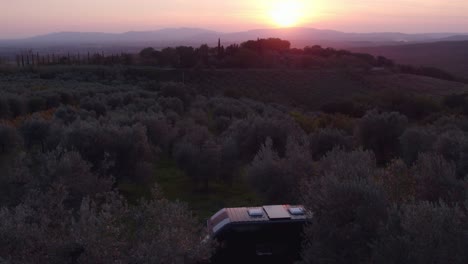 Camper-van-standing-between-olive-tree-plantation-during-Italian-sunset,-aerial