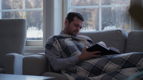 Relaxing-man-with-blanket-reads-book-in-couch,-Hygge-concept