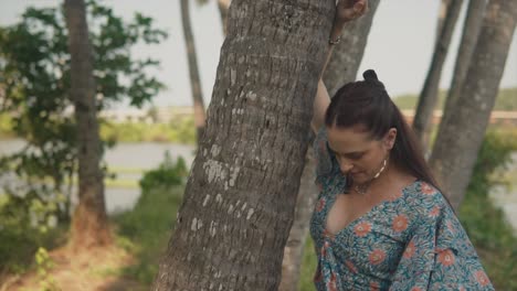 A-close-up-shot-of-a-beautiful-Caucasian-female-in-a-floral-dress-resting-on-the-trunk-of-a-large-palm-tree,-looking-down-at-the-ground-inquisitively