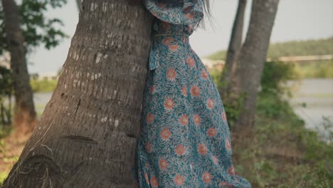 A-pedestal-shot-following-the-trunk-of-a-palm-tree,-a-carefree-female-wearing-a-pretty-blue-floral-dress-embraces-the-tree-showing-her-love-for-nature