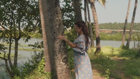 Young-woman-in-a-blue-dress-standing-among-tress-in-a-forest-near-a-lake-smiling