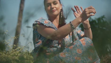 A-close-up-shot-of-an-attractive-Caucasian-female-in-a-beautiful-summers-dress,-pushing-her-hair-back-as-she-poses-and-smiles-for-the-camera-while-sitting-in-a-grass-field-outdoors,-India