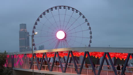 Nächtlicher-Blick-Auf-Eine-Fußgängerbrücke,-Die-Mit-Chinesischen-Roten-Laternen-Geschmückt-Ist,-Während-Die-Menschen-Hindurchgehen,-Während-Sich-Im-Hintergrund-Ein-Riesenrad-In-Hongkong-Befindet