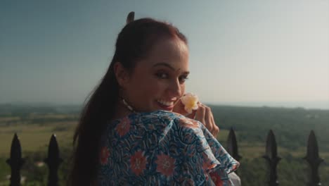 A-close-up-of-a-Caucasian-female-wearing-a-beautiful-summers-dress-holding-a-flower,-looking-back-over-her-shoulder-dollying-out-revealing-the-beautiful-natural-surrounding-view