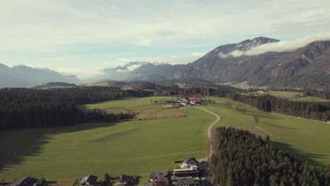 Aerial-drone-of-beautiful-alpine-mountain-range-in-the-distance-with-picturesque-grassy-fields,-trees-and-a-town