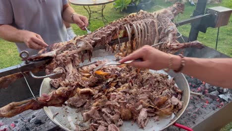 Manos-De-Hombres-Cortando-Un-Cordero-Entero-Con-Tenedores-Como-Salvajes-En-Un-Palo-De-Metal-Escupido-Con-Piel-Dorada-Crujiente,-Barbacoa-De-Cordero-Fiesta-En-El-Jardín-De-Verano,-Chicos-Cocinando,-Tiro-De-4k