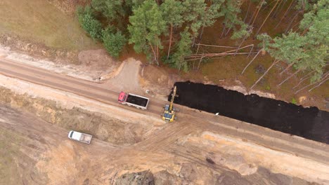 Yellow-digger-and-dump-truck-working-on-road-construction,-aerial-top-down-orbit-view