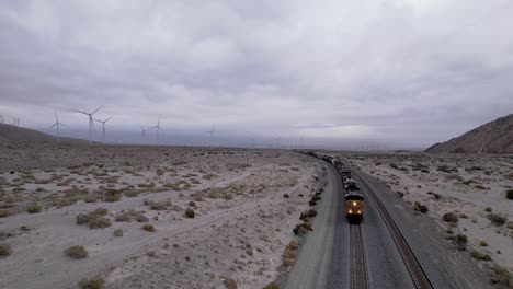 Cargo-train-passing-by-the-desert-with-wind-turbines,-USA