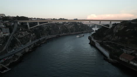 Sunset-Drone-Shot-Following-Boat-Underneath-Massive-Bridge-in-Porto,-Portugal