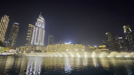 Asombroso-Espectáculo-De-Agua-Desde-La-Fuente-De-Dubai-Visto-De-Noche-Con-El-Paisaje-Urbano-Al-Fondo