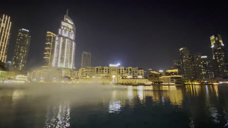Rauch-Auf-Dem-Wasser-Im-Dubai-brunnen,-Der-Nachts-Mit-Luxuriösen-Restaurants-Im-Hintergrund-Zu-Sehen-Ist