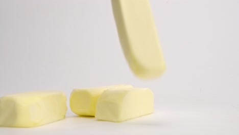 Whole,-unwrapped-sticks-of-yellow-butter-cubes-falling-and-bouncing-onto-white-table-top-in-slow-motion-and-landing-in-pile