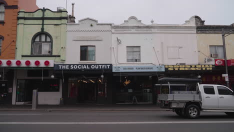 Cars-passing-row-of-shops-in-the-suburb-of-Newtown,-Sydney,-Australia-on-gloomy-overcast-rainy-day