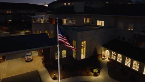 Aerial-orbit-around-American-flag-waving-in-front-of-retirement-home-building-at-night